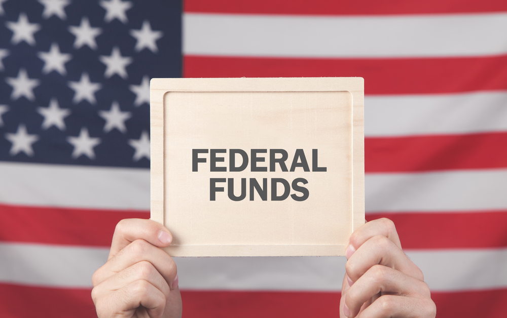 Hands hold a sign that reads “Federal Funds” in front of U.S. flag.