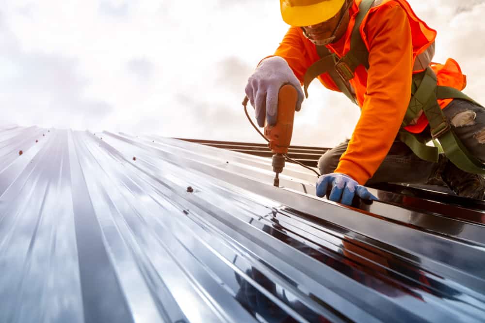 Image of a man in a hard hat using a drill to nstall solar panels on a metal roof.